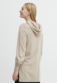 Haut à capuche beige à manches longues avec une coupe décontractée, en tissu tricoté texturé, avec capuche et poignets retroussés, s'étendant sous les hanches.