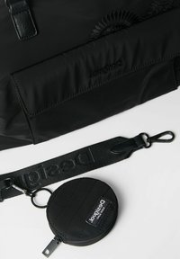 Borsa in tessuto nero con una tracolla removibile e un sacchetto rotondo con cerniera, entrambi etichettati "Desigual," posati su una superficie bianca.