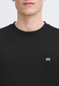 Tricou din bumbac negru cu guler tip crew și un logo brodat alb pe partea stângă a pieptului. Textură netedă, design minimalist.