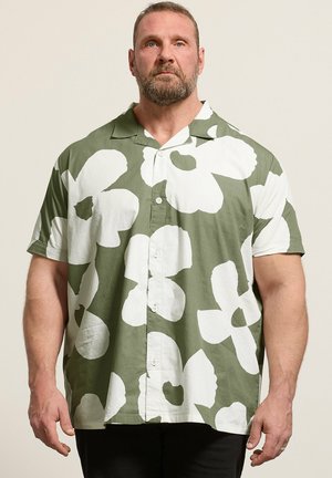 Mann mit Bart, der ein grünes kurzärmeliges Hemd mit großem weißem Blumenmuster trägt, steht vor einem schlichten hellen Hintergrund.