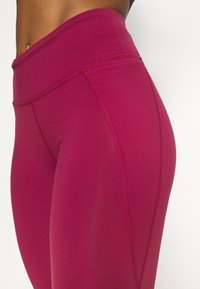 Höga, åtsittande leggings i en djup magenta färg, tillverkade av stretchigt material med synlig sömdetaljering längs sidorna.