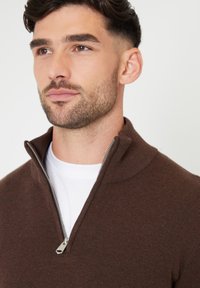 Brauner Strickpullover mit einem halben Kragen und Reißverschluss sowie einem gerippten Muster, über einem weißen Hemd getragen. Mit einem silbernen Reißverschlussdetail.