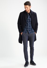 Dark coat over a green and navy plaid shirt, paired with textured navy pants and dark shoes. The outfit features a tailored fit and distinct patterns.