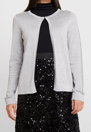 Femme portant un cardigan scintillant gris clair par-dessus un col roulé noir et une jupe noire à sequins sur un fond uni.