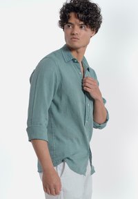 Lichtgroene linnen shirt met lange mouwen, opgerold tot aan de ellebogen, met een geknoopte kraag en een afgeronde zoom, gecombineerd met witte broek.