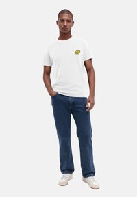 Weißes Baumwoll-T-Shirt mit einem gelben Zitronenmotiv auf der linken Brust, kombiniert mit blauen Jeans und weißen Sneakers.
