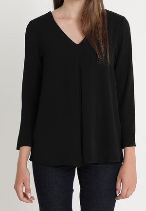 Blouse noire à manches longues avec un décolleté en V, en tissu lisse. Présente une coupe décontractée et des coutures décoratives descendant sur le devant.