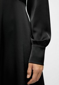 Højre hånd afslappet ved siden iført en lang sort kjole med tætsiddende manchette og let sammensnøring af stoffet over håndleddet.