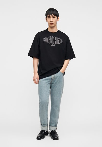 Μαύρο υπερμέγεθες μπλουζάκι με λευκό λογότυπο "MOSCHINO", σε συνδυασμό με τζιν παντελόνι ανοιχτού μπλε χρώματος και μαύρα μοκασίνια, στέκεται μπροστά σε απλό φόντο.