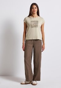 Světle béžové bavlněné tričko se čtvercovou grafikou "TICHÉ POČITKY," v kombinaci s volnými hnědými lněnými kalhotami a béžovými teniskami.