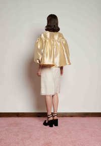 Sieviete, vērsta pret sienu, valkājot spīdīgu zeltainu pūderētu apmetni, ceļgala garuma krēmkrāsas svārkus ar nelieliem ziedu rakstiem un melnas sandales ar biezām papēžiem.