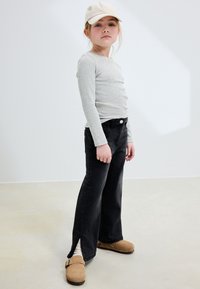 Graues langärmliges Hemd, schwarze ausgestellte Jeans mit seitlichen Schlitzen und beige Clogs. Das Kind trägt eine hellbeige Mütze und hat glattes Haar.