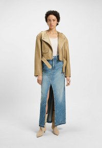 Veste en cuir beige cropped avec des fermetures éclair argentées, associée à une jupe en denim bleu à fente haute avec un design délavé et des talons moyens.