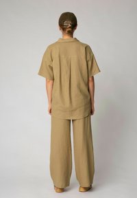 Khakifarbene Leinenbluse mit kurzen Ärmeln und abgerundetem Saum, kombiniert mit weiten Khakileinenhosen. Das Outfit wird durch eine braune Mütze mit Schrift ergänzt.