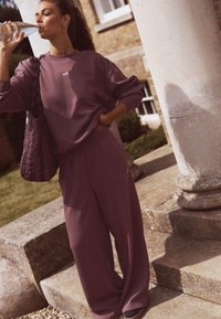 Ljubičasti pamučni džemper s natpisom "self.", uparен s širokim ljubičastim hlačama. Modni dodatak: prošivena torba. Model pije vodu na otvorenom, kraj stupova.