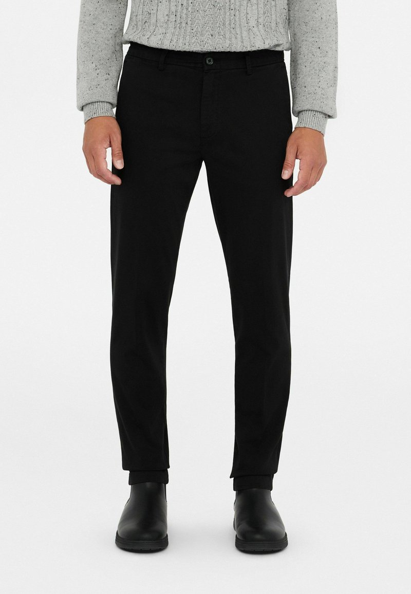 Zwarte trousers met een slim fit, voorzien van een knoopsluiting, riemlussen en een gladde textuur. Gecombineerd met zwarte enkellaarsjes.
