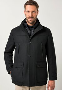 Uomo che indossa un cappotto nero con bottoni, dotato di tasche con zip sul petto e tasche a pattina per le mani, in piedi davanti a uno sfondo chiaro e uniforme.