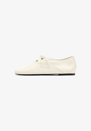 Chaussure plate couleur crème, à lacets devant, œillets dorés et semelle fine noire, montrée en profil gauche sur fond blanc.
