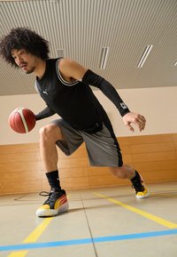 Top sin mangas negro con detalles en blanco, pantalones cortos grises y zapatillas de colores; jugador driblando un balón de baloncesto en el suelo de un gimnasio.