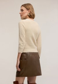 Kobieta w kremowym, puszystym swetrze i brązowej skórzanej spódnicy z zapiętym na guziki kieszonką, nosząca złote i perłowe kolczyki, zwrócona bokiem na beżowym tle.