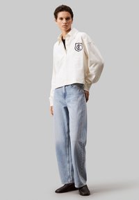 Maglietta a collo bianco con logo ricamato nero, abbinata a jeans a gamba dritta a vita alta di colore blu chiaro e stivali neri alla caviglia.
