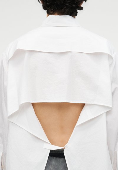 Person mit lockigem Haar, die ein weißes Langarmshirt mit einem dreieckigen, offenen Rückendesign trägt, das nackte Haut zeigt.