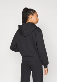 Μαύρη φούτερ ζακέτα με άνετη εφαρμογή, κουκούλα με κορδόνι και ριμπ μανσέτες. Το ύφασμα φαίνεται μαλακό και υφή, συμπληρωμένο με αντίστοιχο μαύρο φόρμα παντελόνι.