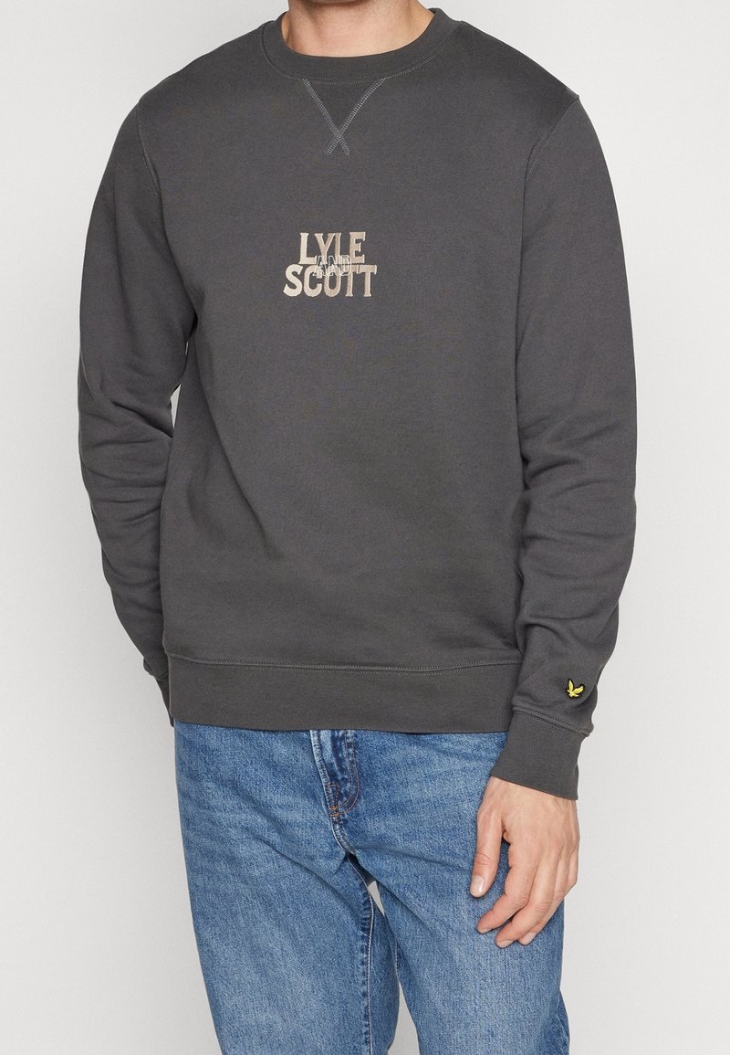 Homme portant un sweat-shirt gris foncé à col rond avec le texte "Lyle and Scott" et un logo d'oiseau jaune sur le poignet, assorti à un jean bleu.