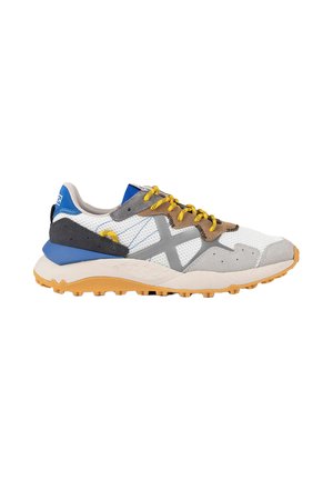 Zapatilla blanca y gris con talón azul, cordones amarillos, detalles en color tostado y suela de goma texturizada, mostrada de lado sobre un fondo blanco.
