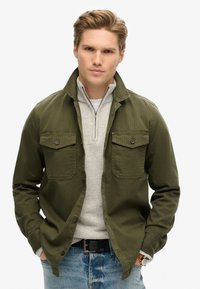 Chaqueta de botón verde oliva con dos bolsillos en el pecho, llevada sobre un suéter gris de cremallera, combinada con jeans azul claro y un cinturón negro.