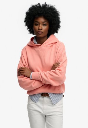 Femme aux cheveux bouclés portant un sweat à capuche rose et une chemise rayée, les bras croisés, debout devant un fond blanc uni.