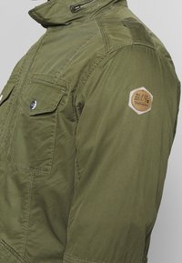 Veste vert olive avec un col élevé, fermeture éclair à l'avant, poches à rabat et un patch hexagonal de marque sur la manche gauche. Texture de tissu légère.