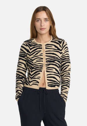 Frau mit hellbraunen Haaren trägt einen beigen und schwarzen zebragestreiften, geknöpften Cardigan und schwarze Kordelhose mit Händen in den Taschen.