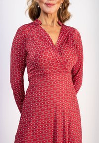 Punainen mekko pitkillä hihoilla, V-pääntiellä ja tiukalla yläosalla. Sisältää toistuvan valkoisen pyörteen kuvion, jossa on pieniä pisteitä ympäriinsä.