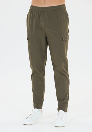 Homme portant un pantalon cargo vert olive avec une taille élastique et des baskets blanches, debout devant un fond blanc uni.