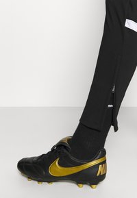 Schwarzer Nike Fußballschuh mit goldenen Akzenten, oberes Material aus strukturiertem Leder und schwarzer Socke. Seitenreißverschluss an schwarzen Sporthosen.