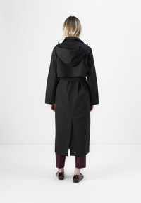 Persona con capelli biondi corti che indossa un lungo trench coat nero, pantaloni croppati borgogna e scarpe marrone scuro, in piedi su uno sfondo bianco.