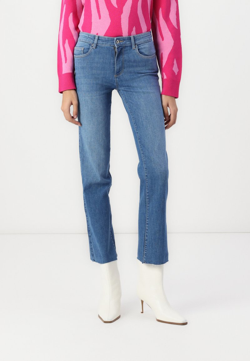 Jeans de mezclilla azul de pierna recta con cinco bolsillos y costuras sutiles, combinados con botas de tobillo blancas con punta y un suéter de diseño rosa.