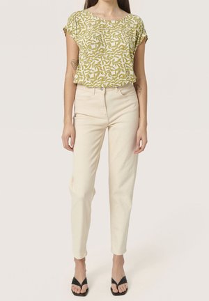 Femme portant un haut à manches courtes à motif de feuilles vertes, un jean taille haute blanc cassé et des sandales à talons noires, debout devant un fond uni.