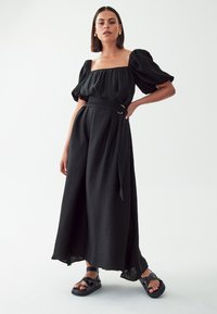 Black dress with puff sleeves, square neckline, and belt detail. Flowing silhouette in textured fabric, paired with black strappy sandals.