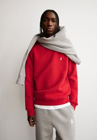 Felpa rossa con un piccolo logo bianco a forma di nastro, abbinata a pantaloni da tuta grigi con tasche laterali. Felpa grigia avvolta sulle spalle.