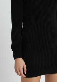 Robe pull noire en tricot avec des manches longues, un tissu texturé à côtes et une silhouette ajustée, présentant un ourlet et des poignets plus bas.
