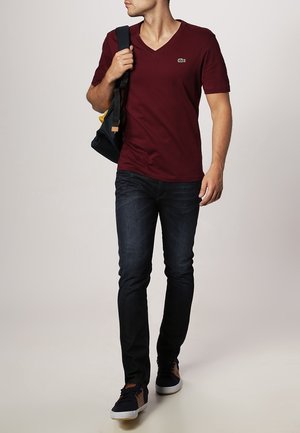 T-shirt bordeaux à col en V, à manches courtes, associé à un jean bleu foncé et des baskets, portant un sac noir et jaune.