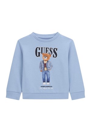 Jasnoniebieska bluza z niedźwiedziem w dżinsowej kurtce i dżinsach, napis "GUESS" u góry oraz "Authentic Denim Brand" na dole.