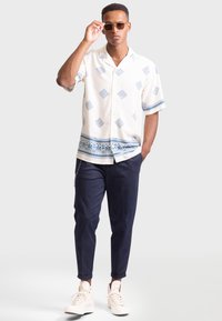 Chemise blanche à manches courtes avec des motifs géométriques bleus, associée à un pantalon navy et des baskets claires, avec un accessoire en chaîne et des lunettes de soleil.