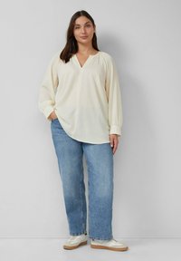 Cremefarbige, strukturierte Bluse mit V-Ausschnitt und gerafften Schultern, kombiniert mit hellblauen Jeans und weißen Sneakers.