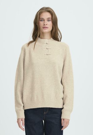 Beige knitted sweater with a high neckline, featuring three toggle buttons and ribbed cuffs and hem, worn over dark jeans.
