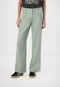 Light green wide-leg trousers with pleats, featuring a smooth texture and a high waistband. Worn with black sneakers and a graphic t-shirt.