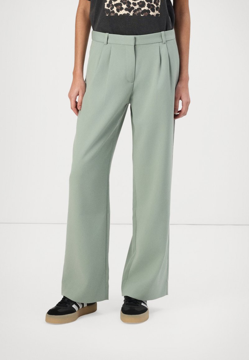 Light green wide-leg trousers with pleats, featuring a smooth texture and a high waistband. Worn with black sneakers and a graphic t-shirt.