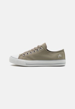 Zapatilla baja de lona beige con puntera de goma blanca, suela blanca con franja gris, ojales metálicos y logo bordado en el talón.